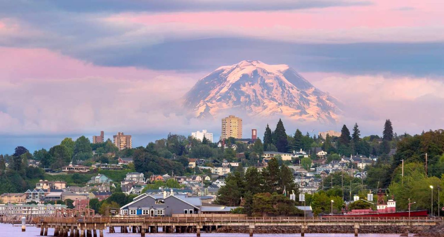 View of a waterfront town with Mount Rainier in the background, under pink and blue skies.