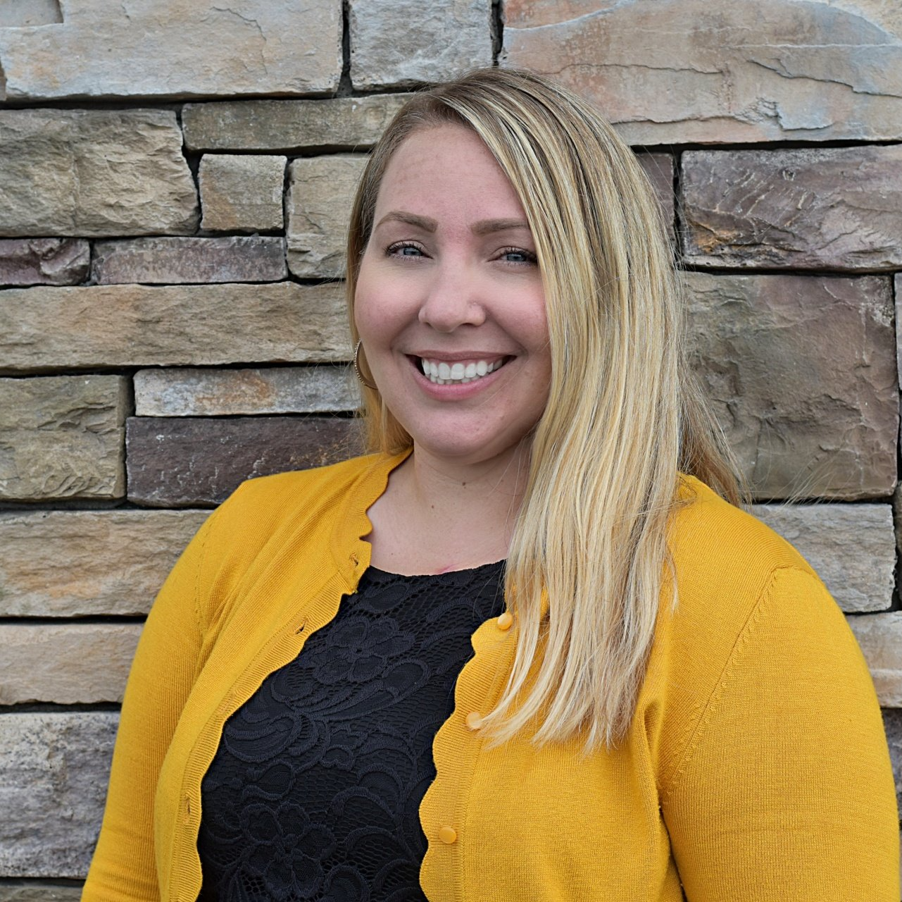 A woman in a yellow cardigan is smiling in front of a stone wall.