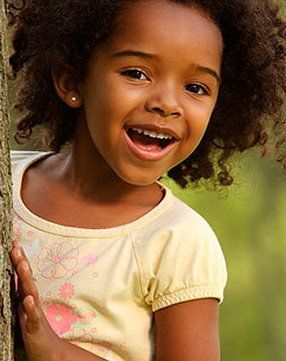 A little girl is peeking out from behind a tree and smiling.