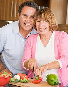 A man and a woman are smiling while cutting vegetables on a cutting board.