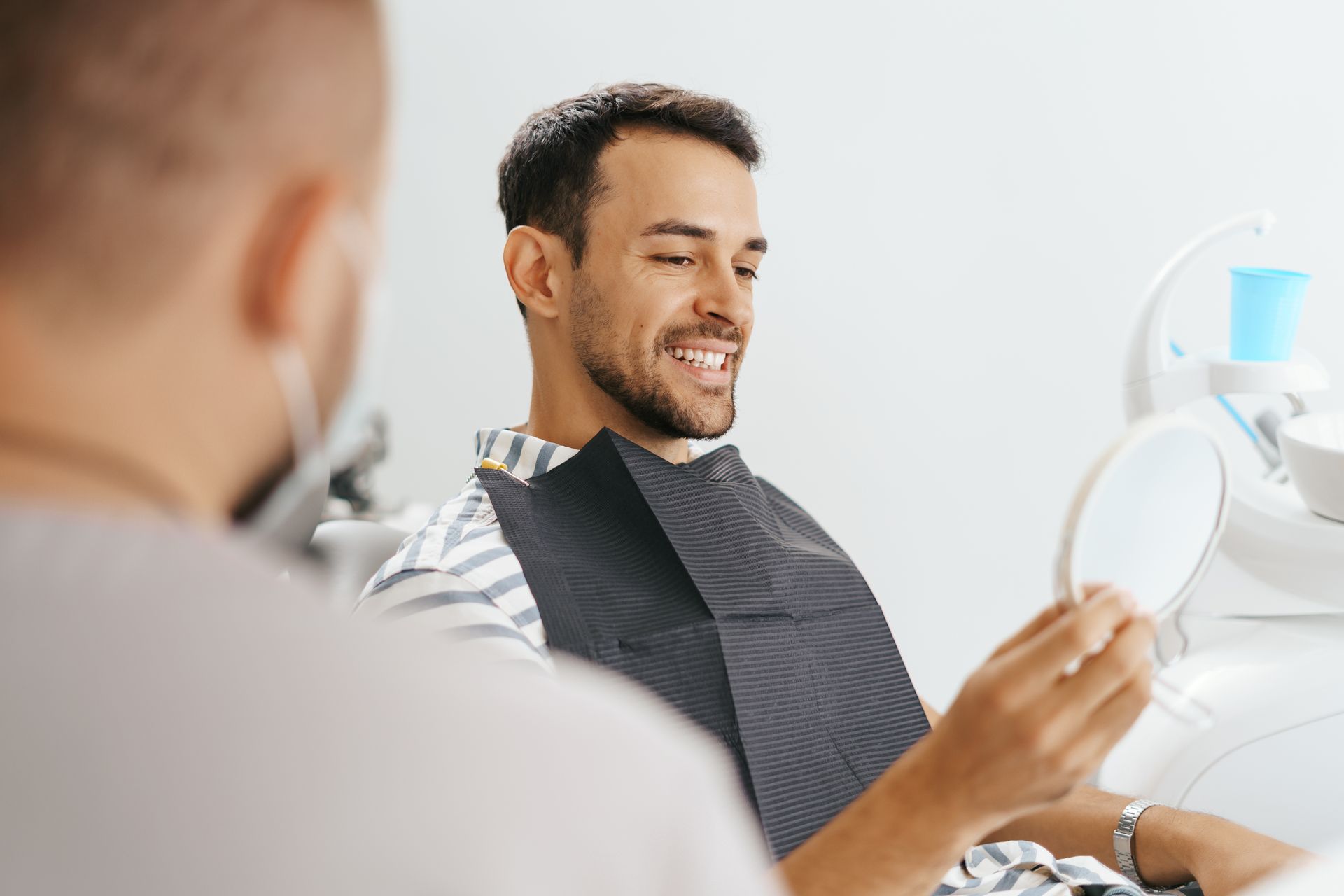 A man is sitting in a dental chair looking at his teeth in a mirror.