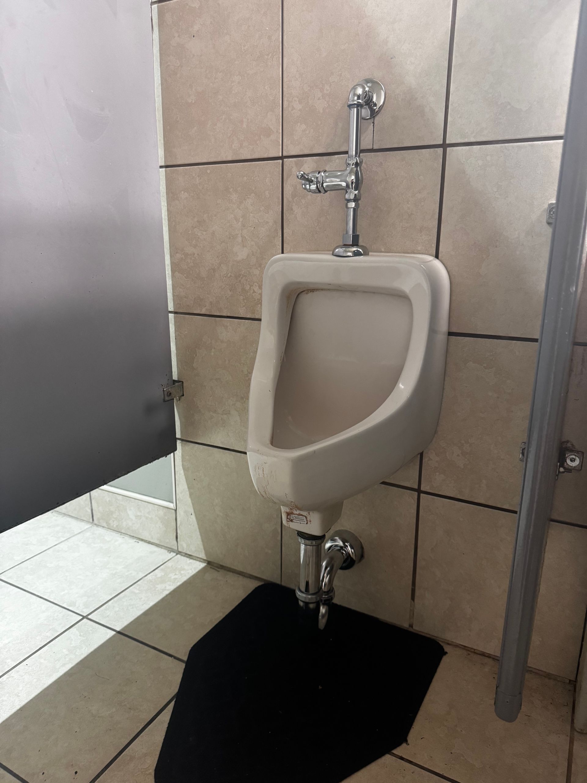 Urinal in a public restroom. White porcelain with chrome fixtures and black floor mat. Beige tiled walls.