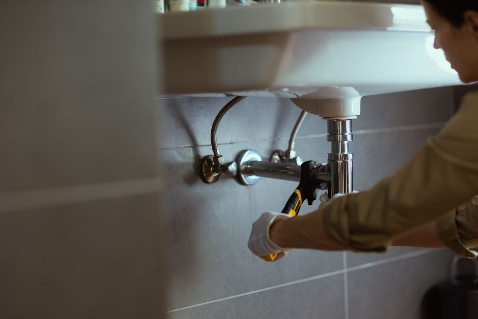 Plumber working under a white sink, using a wrench. Gray tile wall.