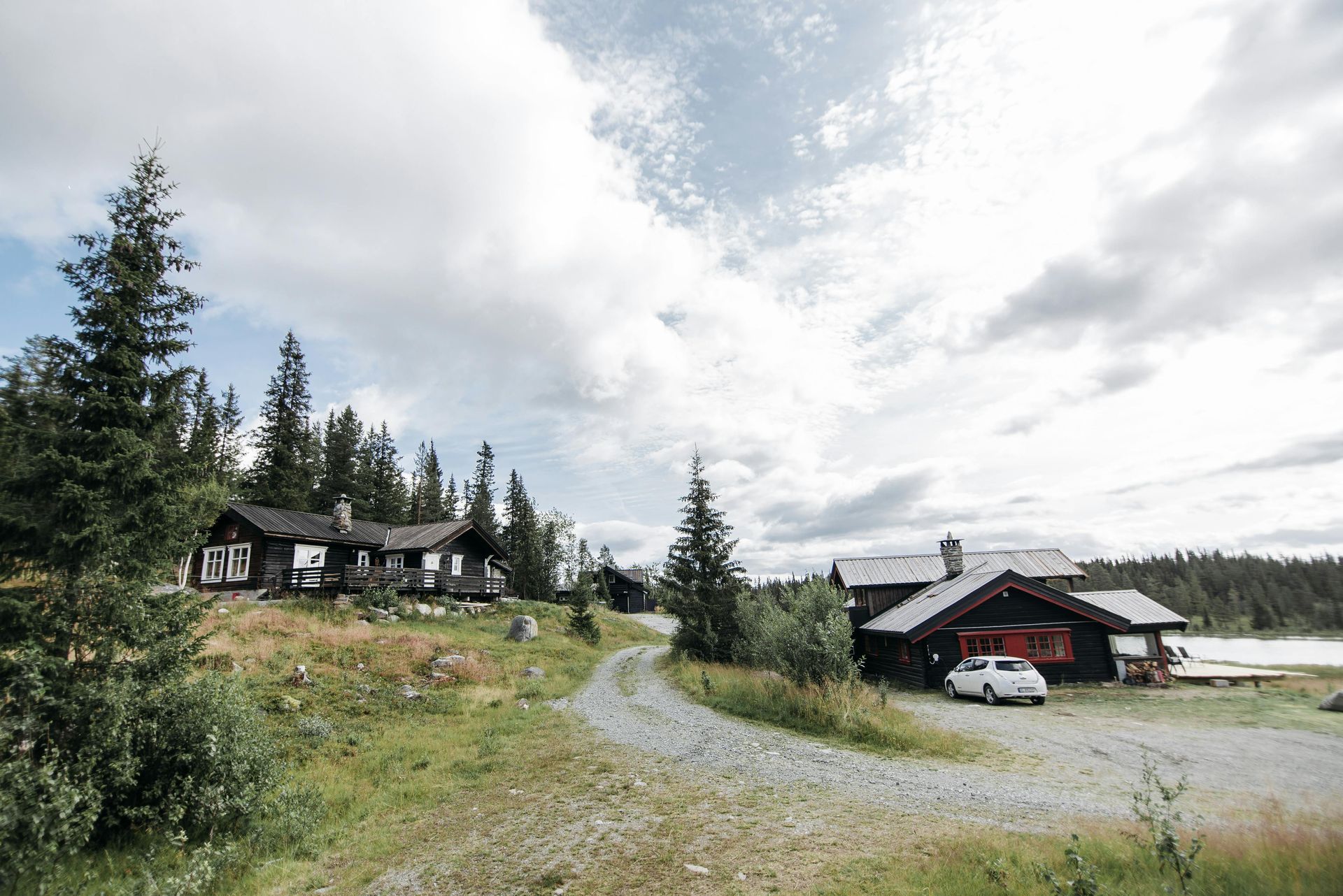 Dirt road leads to wood cabins in a grassy landscape under a cloudy sky.