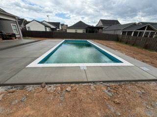 Rectangular backyard swimming pool with a white deck, surrounded by concrete and fencing under cloudy skies
