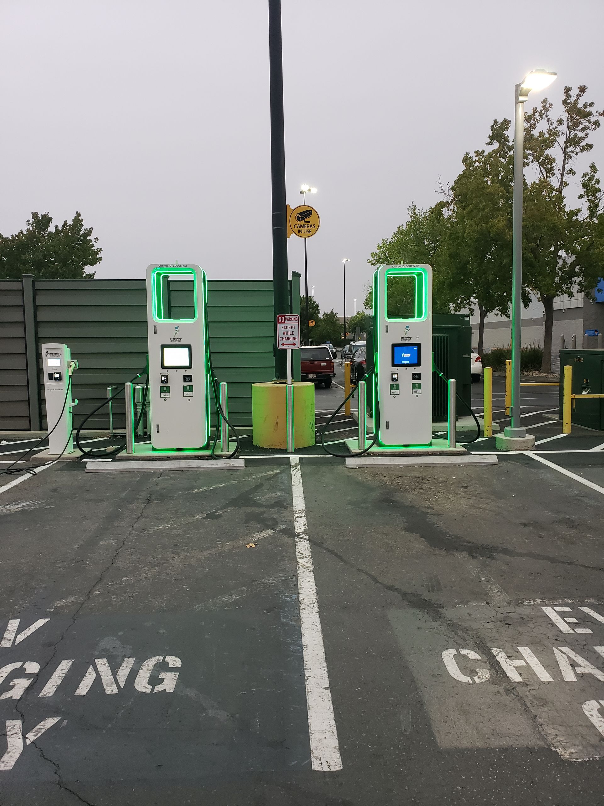 Two green EV charging stations in a parking lot with a central light pole and wet pavement.