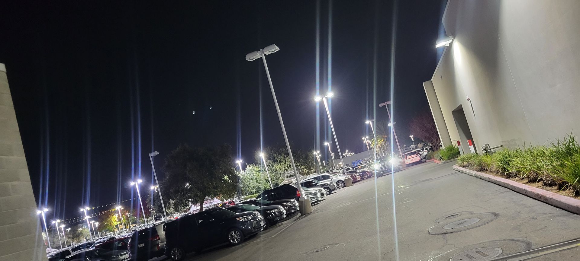 Nighttime parking lot with rows of cars, bright lights, and a walkway beside a building.