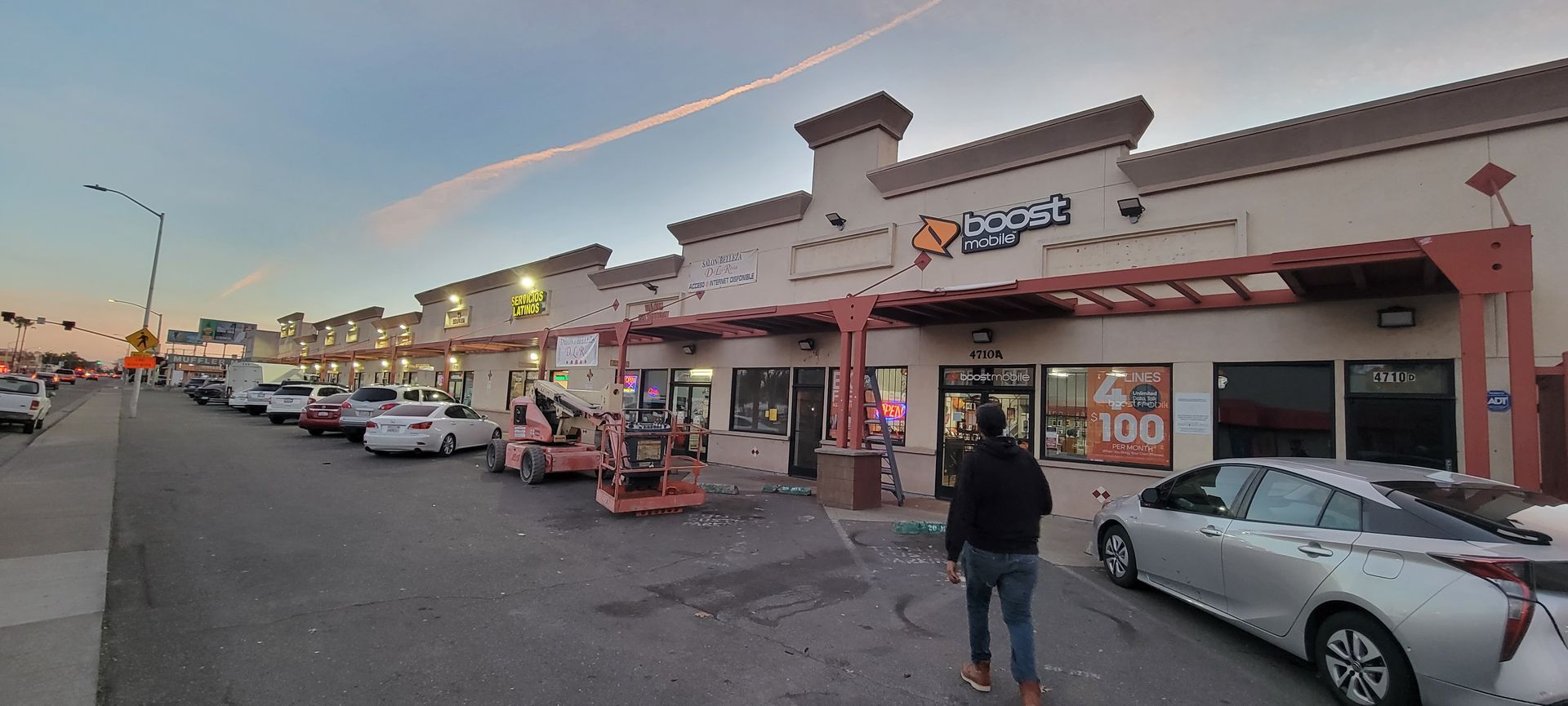 Strip mall storefronts at dusk with parked cars and a person walking along the sidewalk