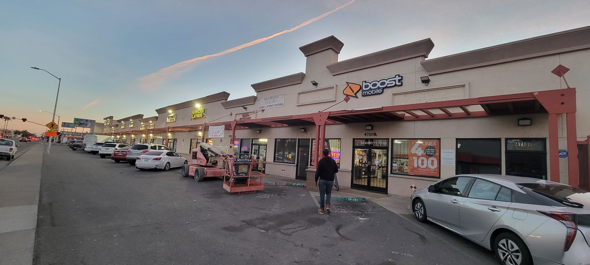Shopping plaza exterior at dusk with storefronts, parked cars, and a few pedestrians in the lot