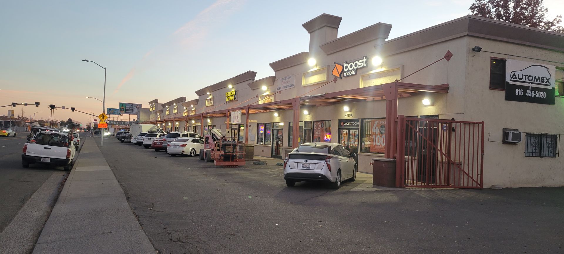 Sunset view of a strip mall with parked cars, lit storefronts, and a sidewalk along the street.