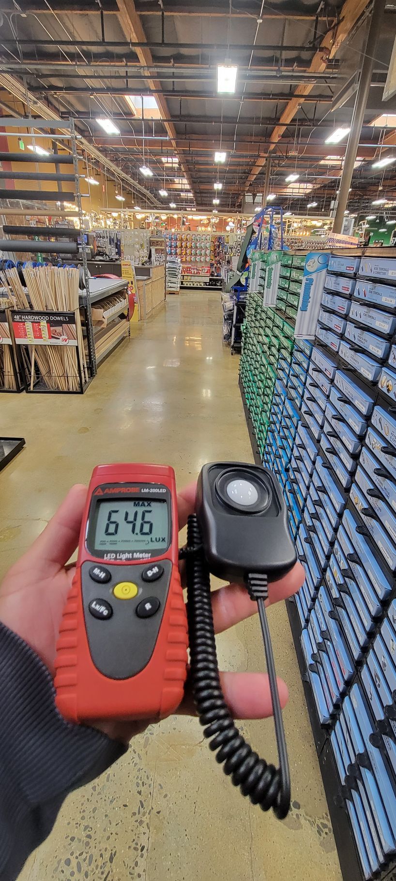 Handheld orange meter with black probe in a warehouse aisle, shelves stacked with boxes on both sides.