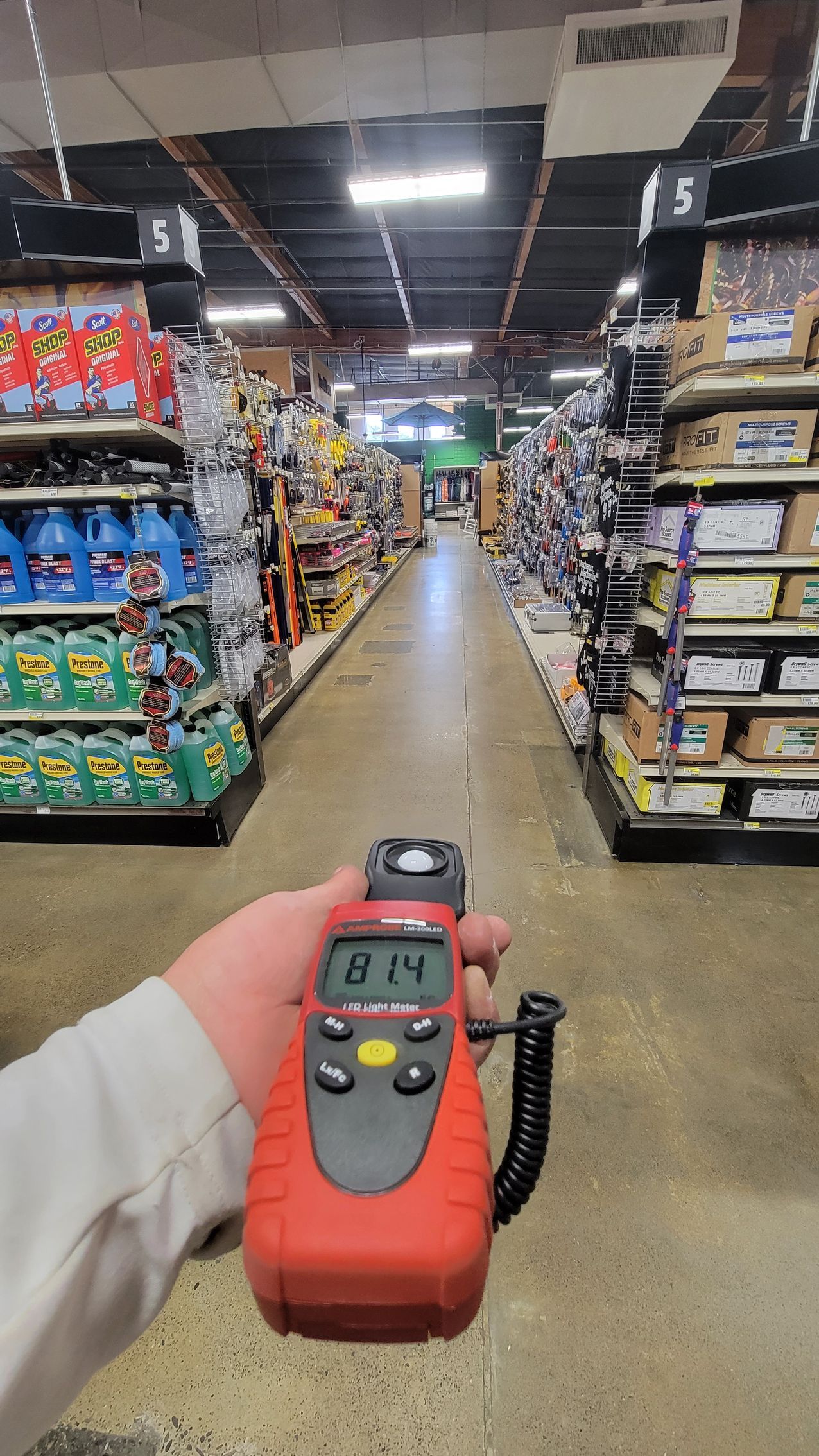 Hand holding a red handheld meter in a grocery store aisle with shelves on both sides.
