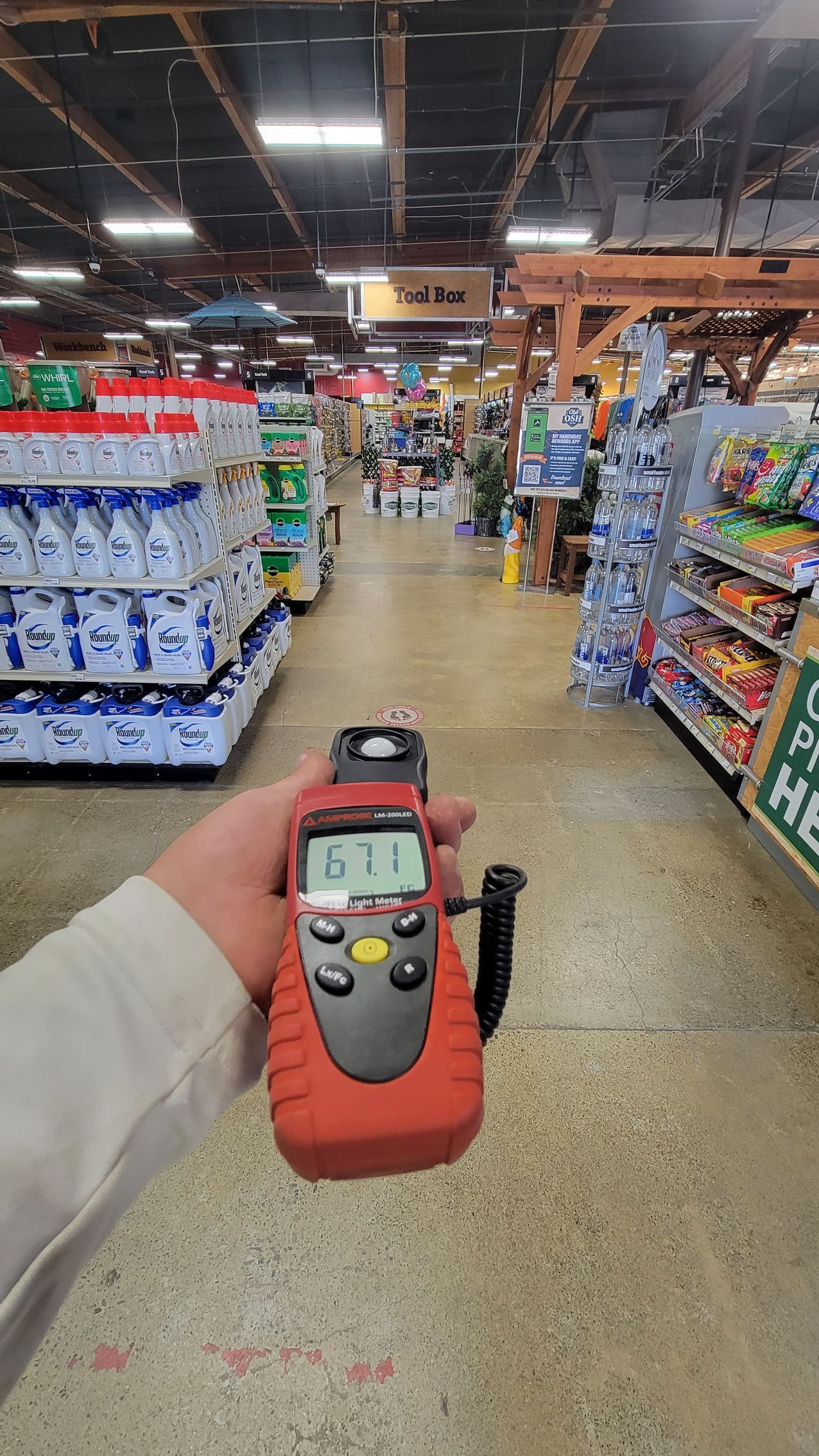 Hand holding a red handheld device in a grocery store aisle with shelves of merchandise.