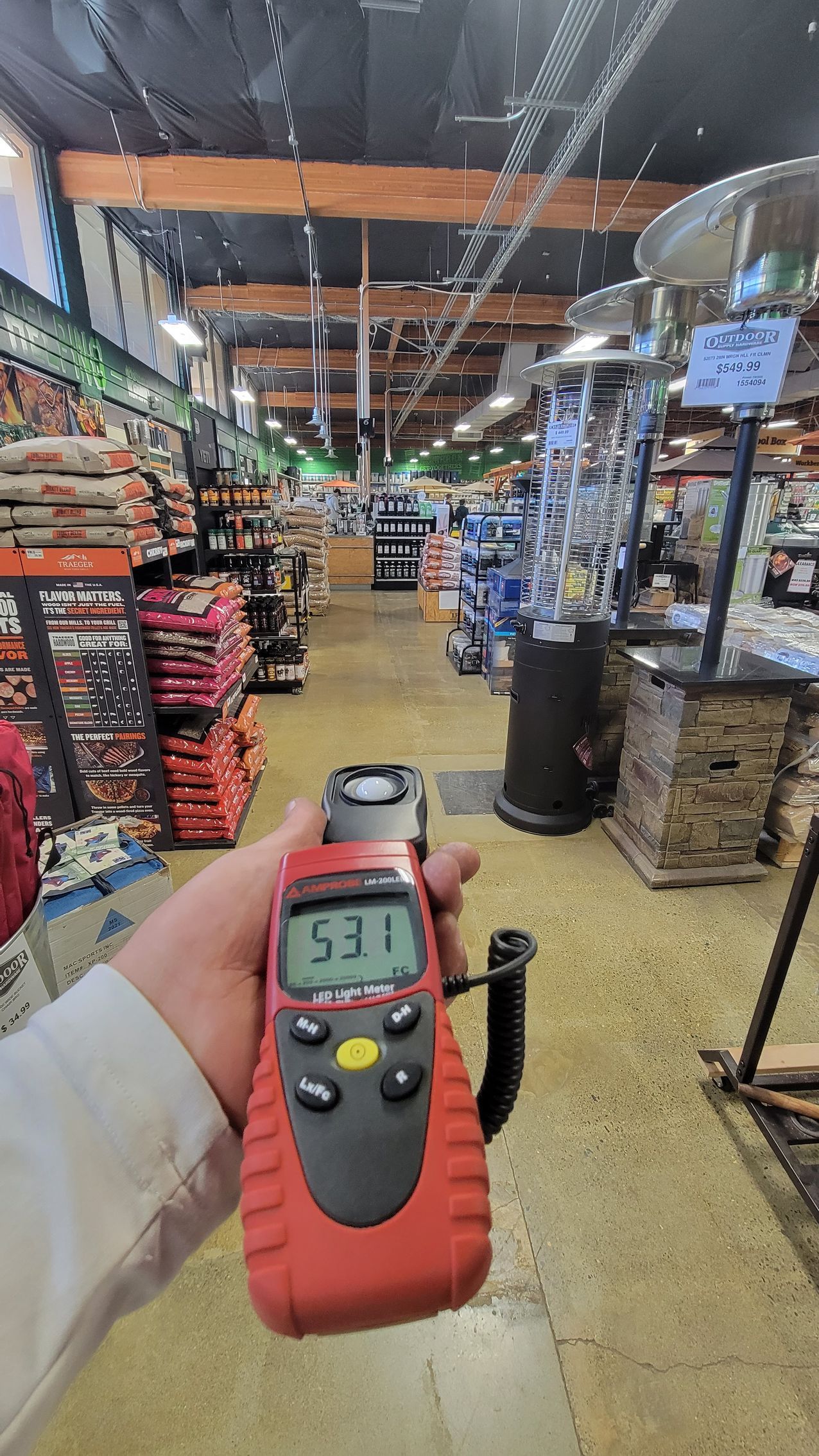 Hand holding a digital meter in a store aisle, with shelves of products and ceiling lights overhead.