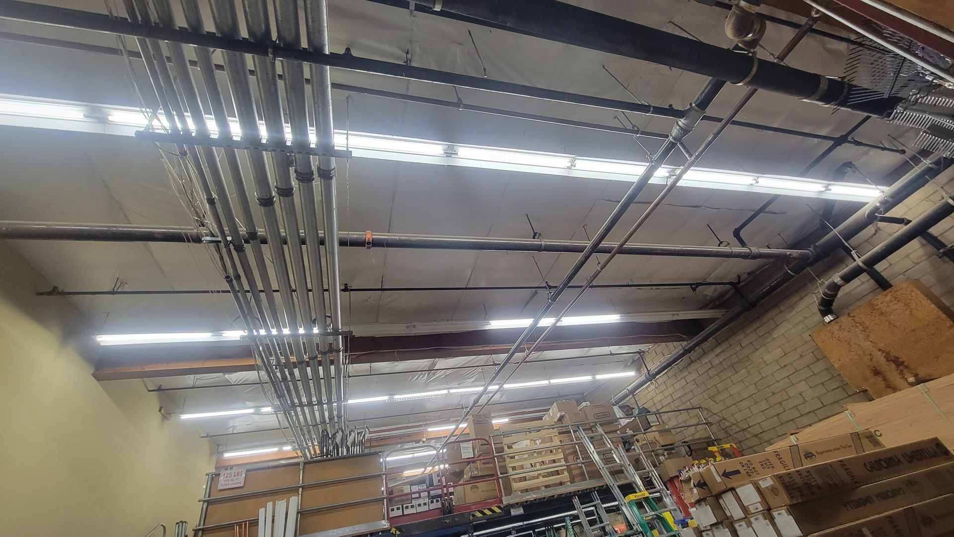 Warehouse ceiling with fluorescent lights, exposed pipes, and shelving below
