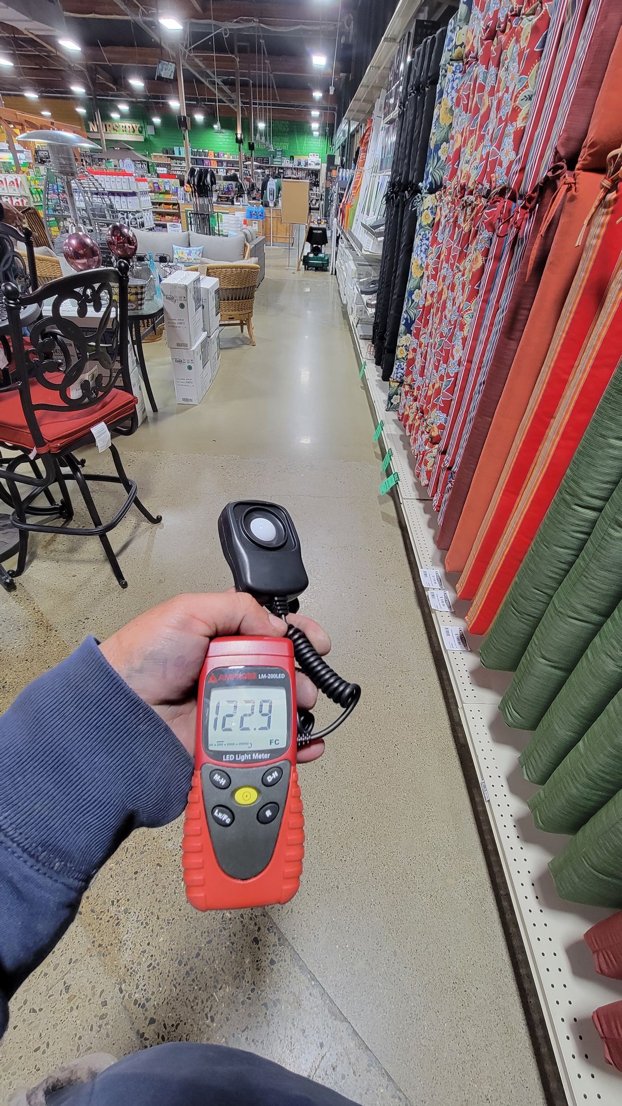 Handheld measuring device in a garden center aisle beside rows of hanging plants and display chairs