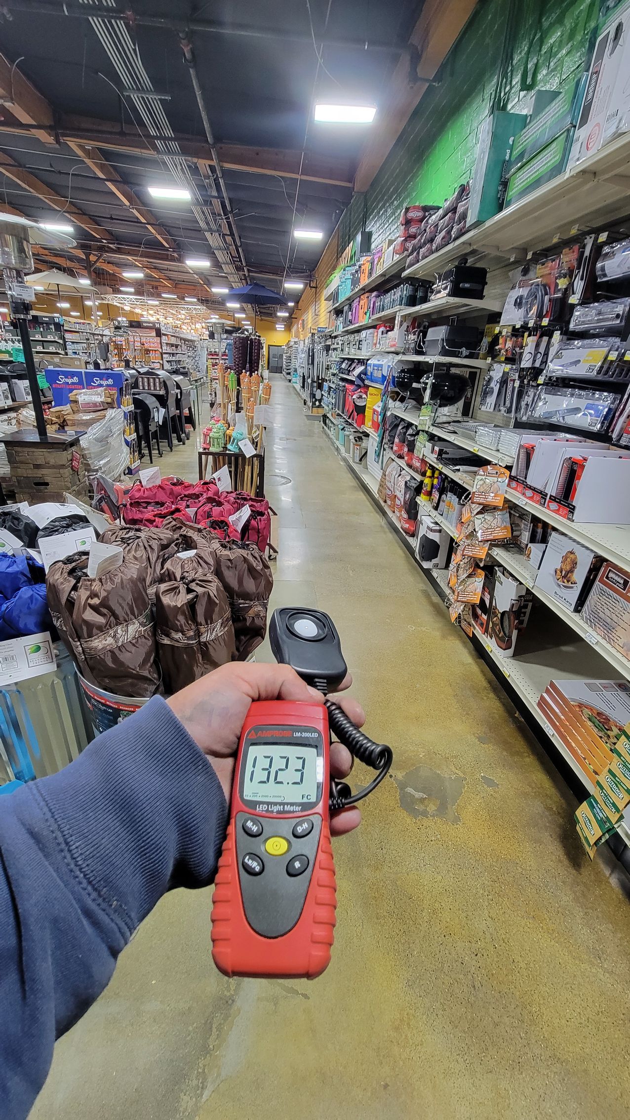 A hand holds a digital meter in a brightly lit hardware store aisle with shelves of tools and supplies.
