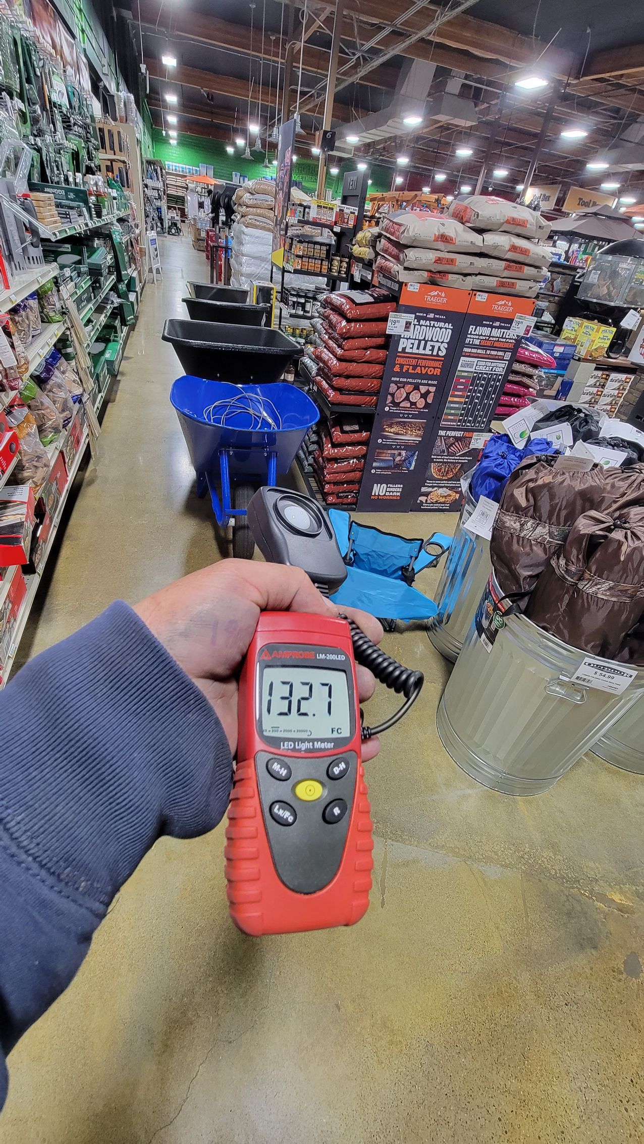 Hand holding a digital meter in a busy warehouse aisle with stacked goods and carts.