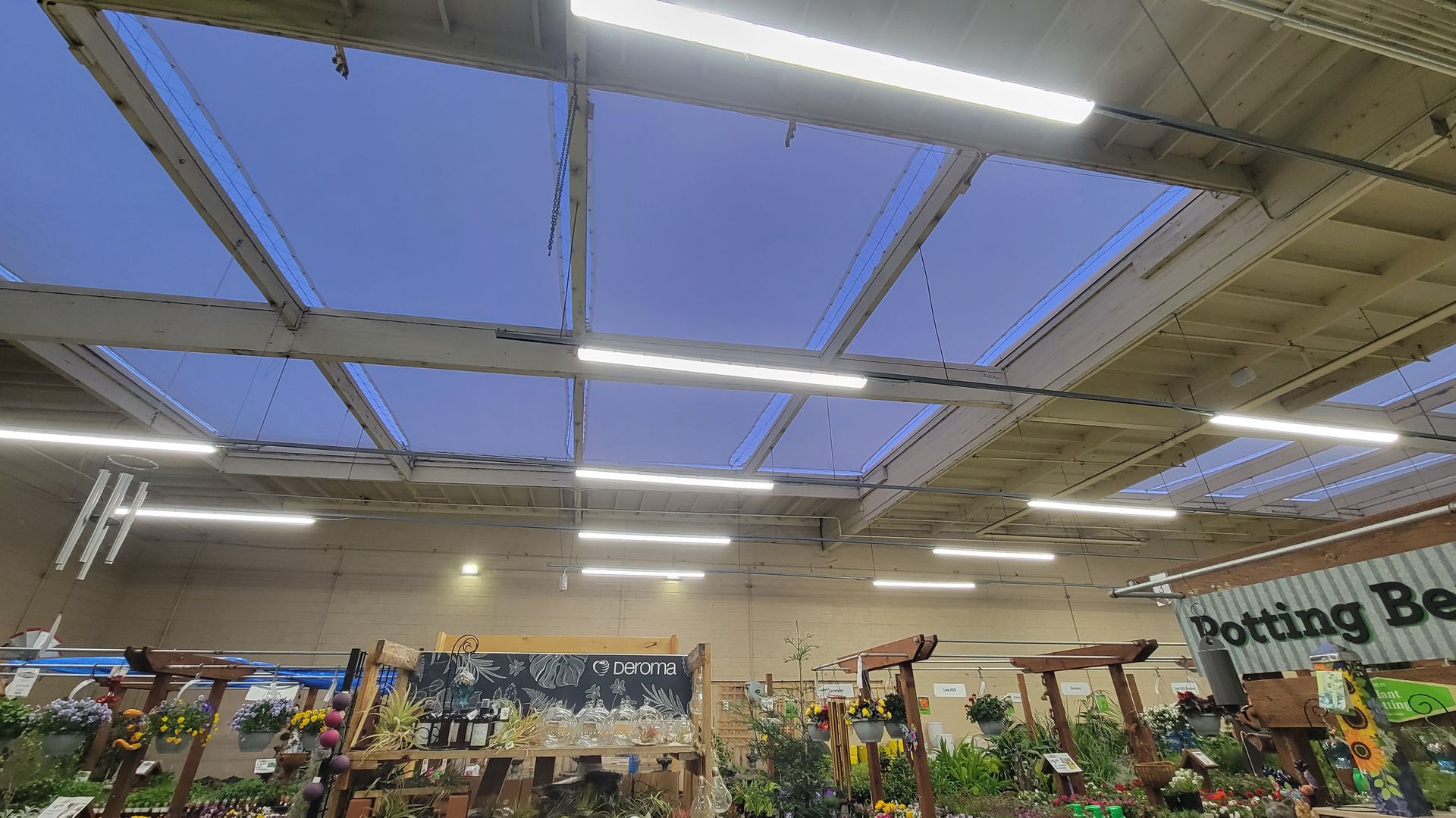 Garden center aisle under bright skylights with rows of plants and a “Potting Bench” sign.