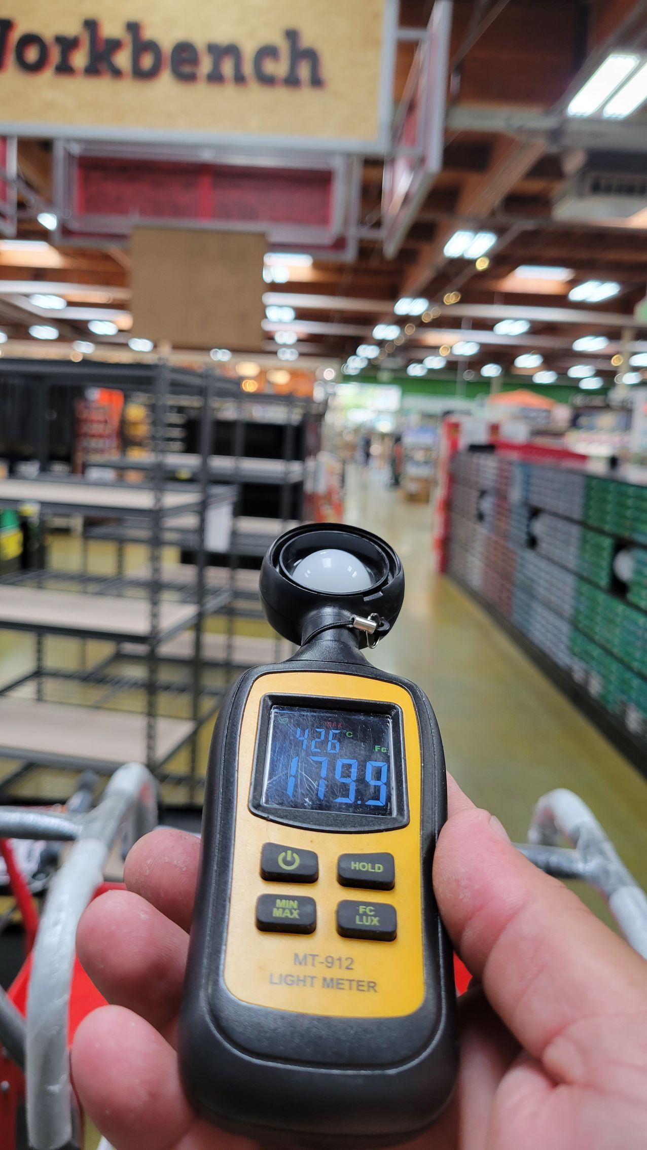 Handheld meter in a store aisle, held up toward shelves and a workbench sign in the background