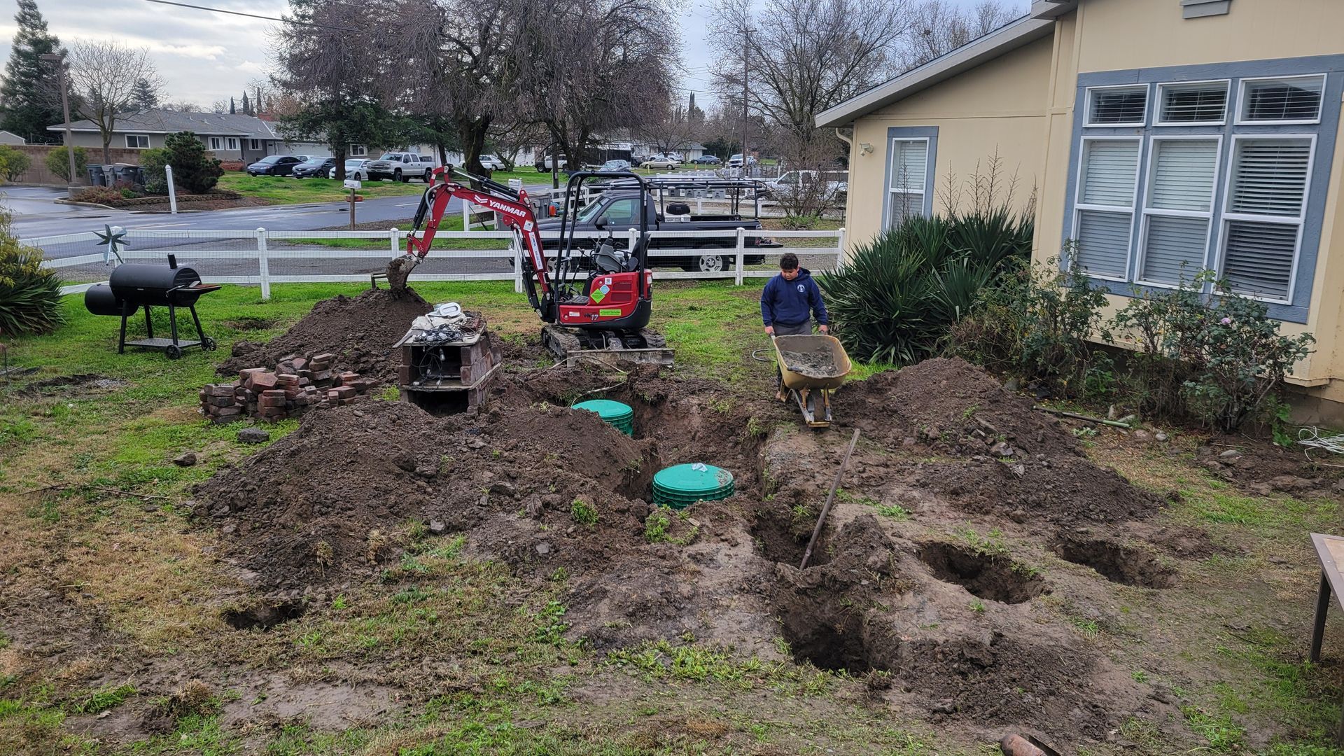 Garden beds under construction beside a house, with muddy soil, tools, and a small excavator.