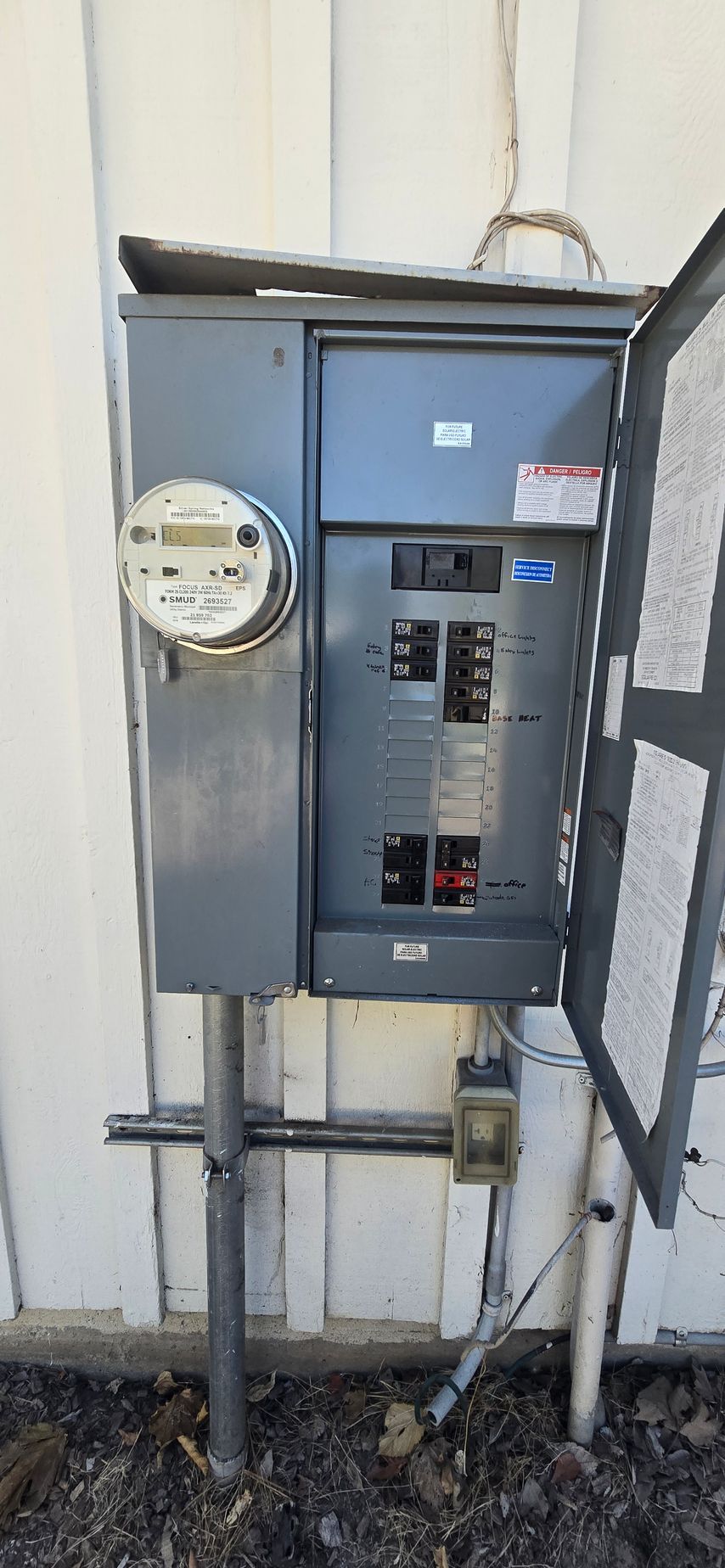 Outdoor electrical meter and breaker panel mounted on a wall, with pipes and gravel below