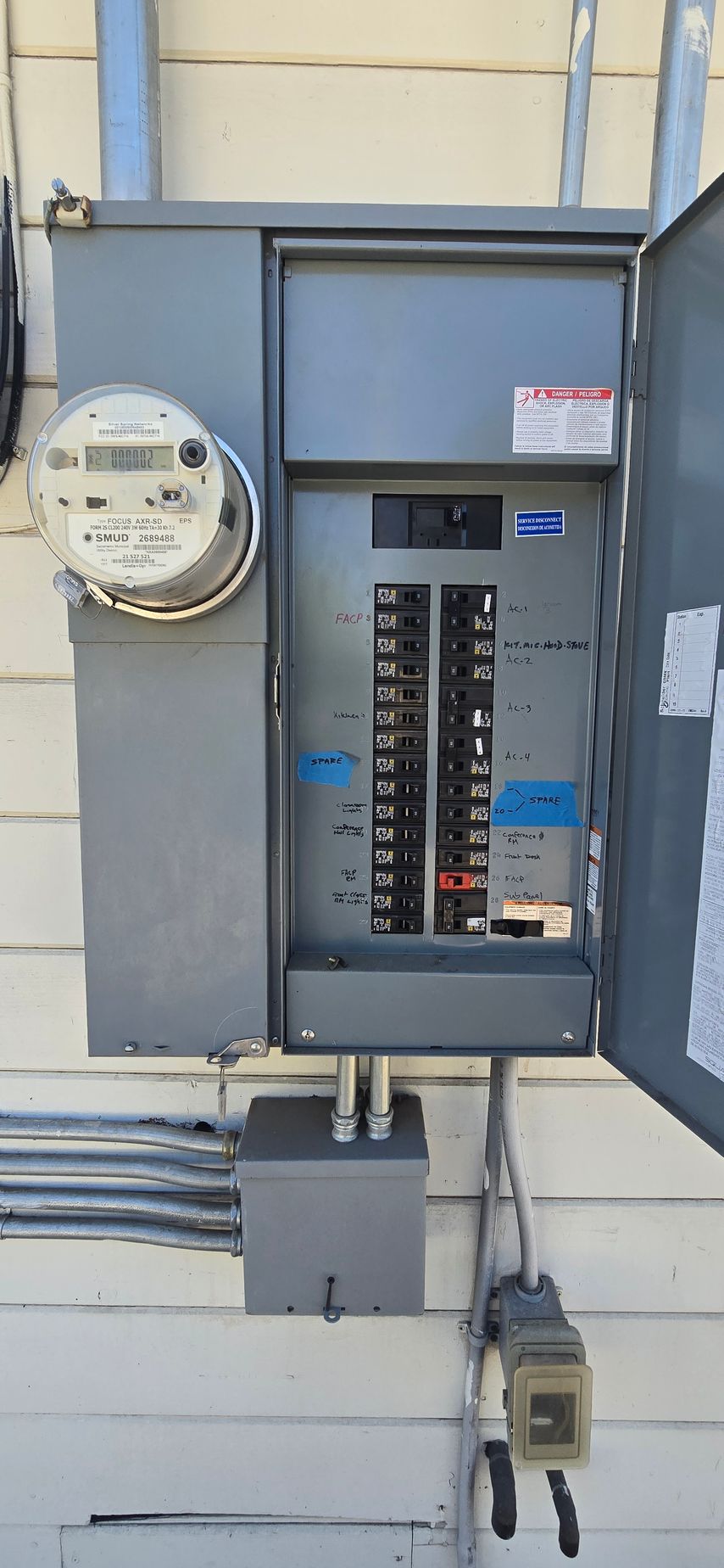 Open electrical breaker panel on a wall with utility meter and conduit pipes.