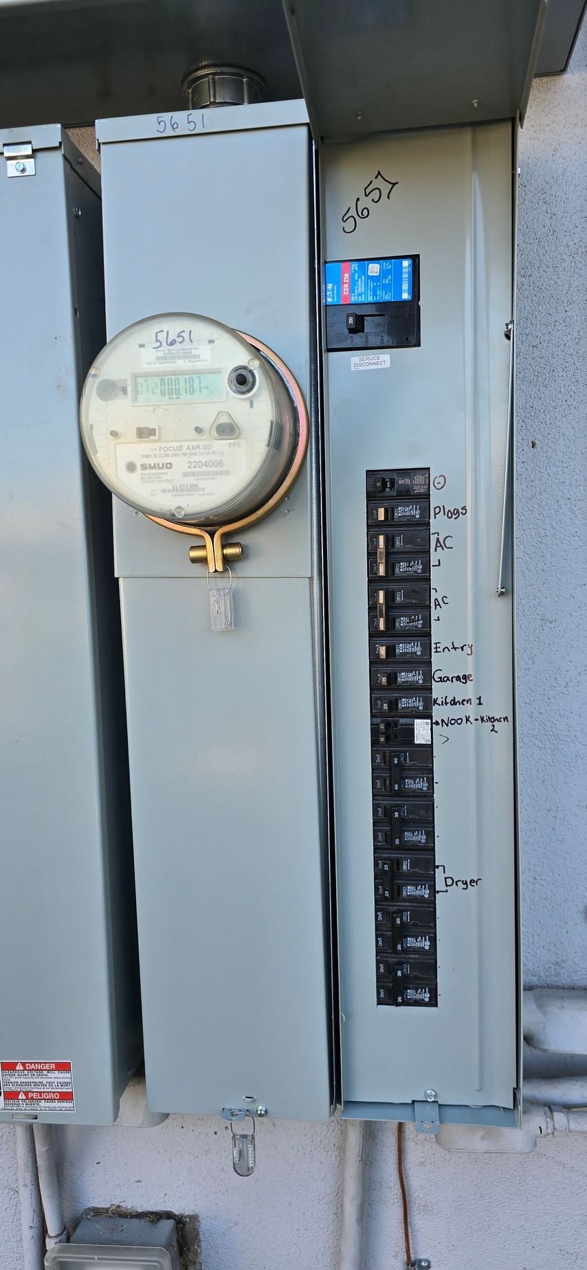 Wall-mounted electric meter and breaker panel on a beige utility box with labels and a blue digital display.