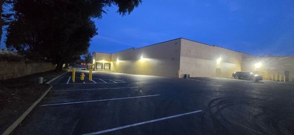 Large warehouse at dusk with lights on, empty parking lot, and trees silhouetted on the left.