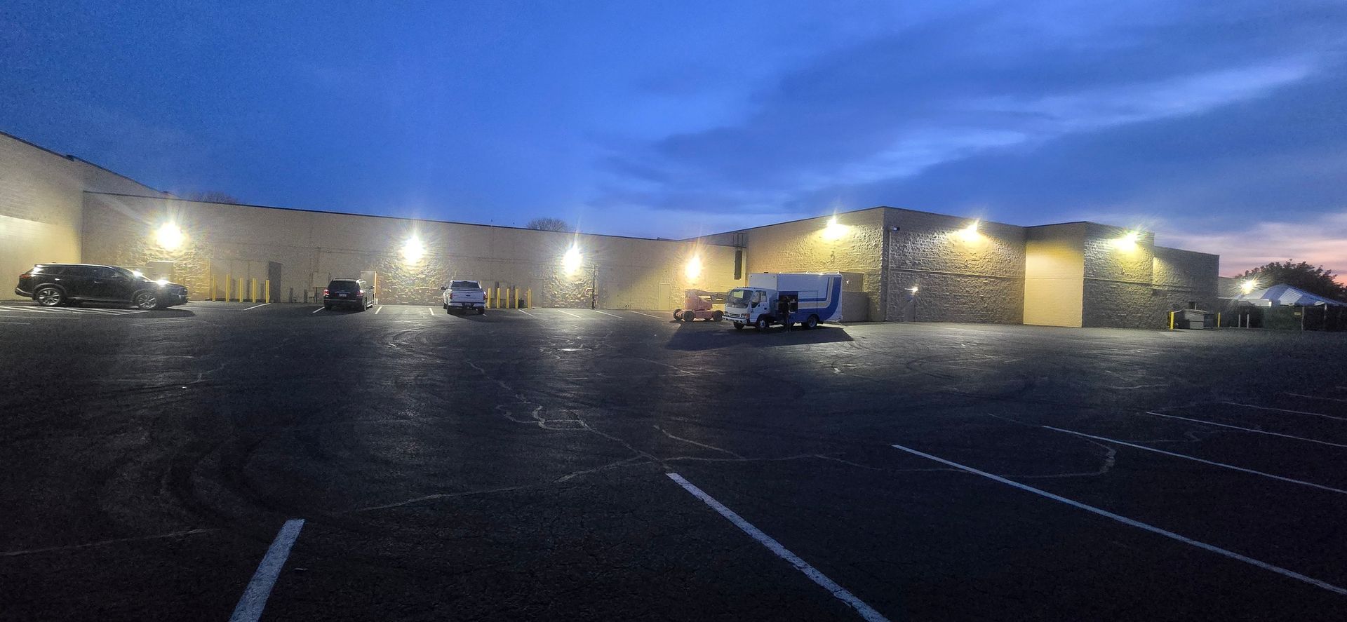 Dimly lit parking lot in front of a low building at dusk, with a few cars and empty spaces.