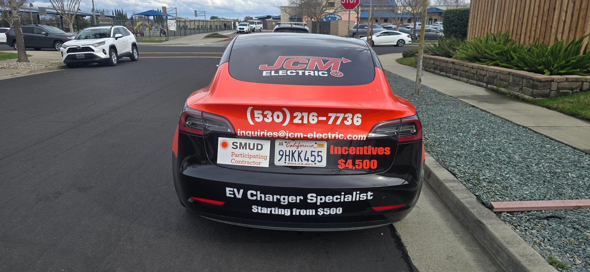 Red-and-black service car with EV charger advertising parked roadside near a curb and gravel landscaping.