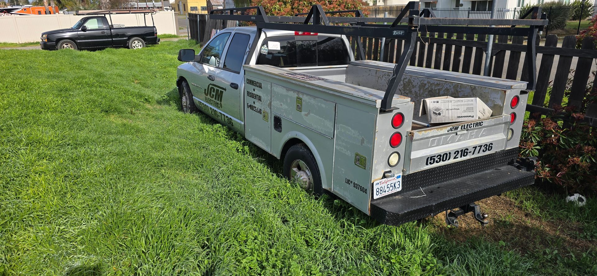 Gray utility pickup truck parked on grass with stacked materials in the bed, near a fence and another truck