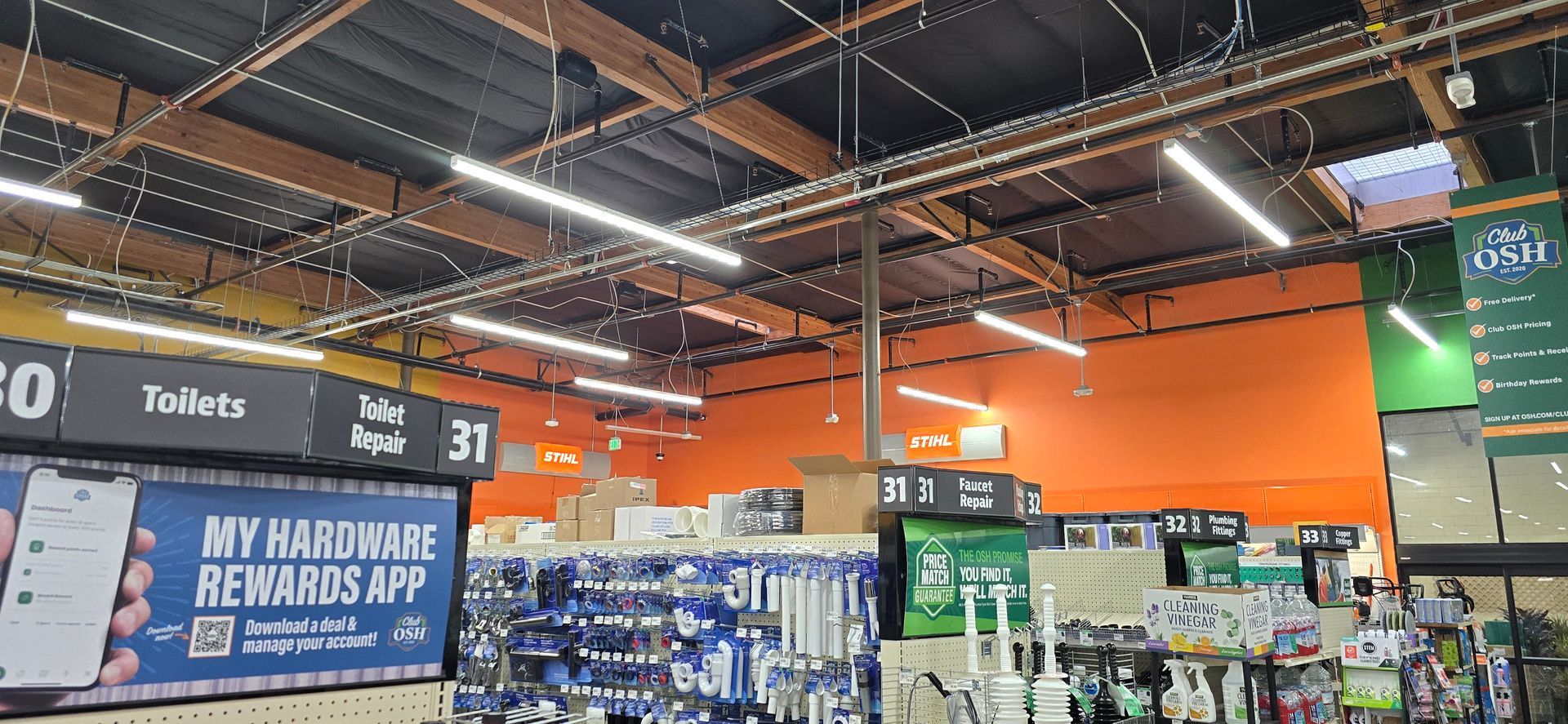 Hardware store aisle with orange ceiling beams, hanging signs, and stacked merchandise displays