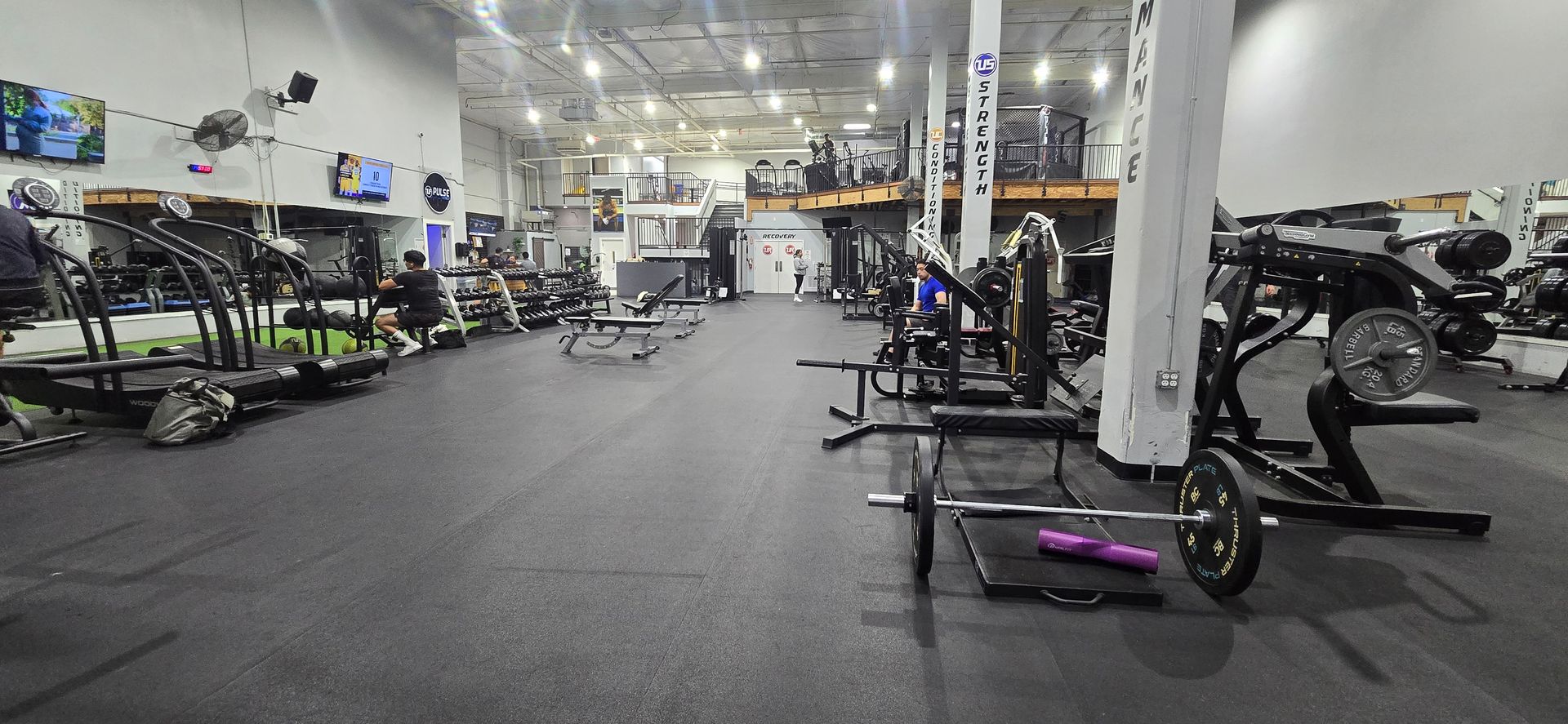 Wide gym interior with weight machines, benches, and dumbbells on a rubber floor