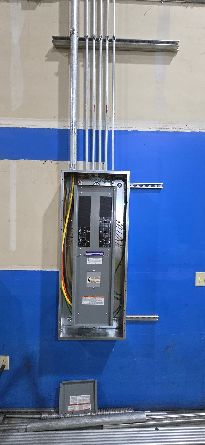 Gray electrical panel mounted on a blue warehouse wall with conduit pipes above it