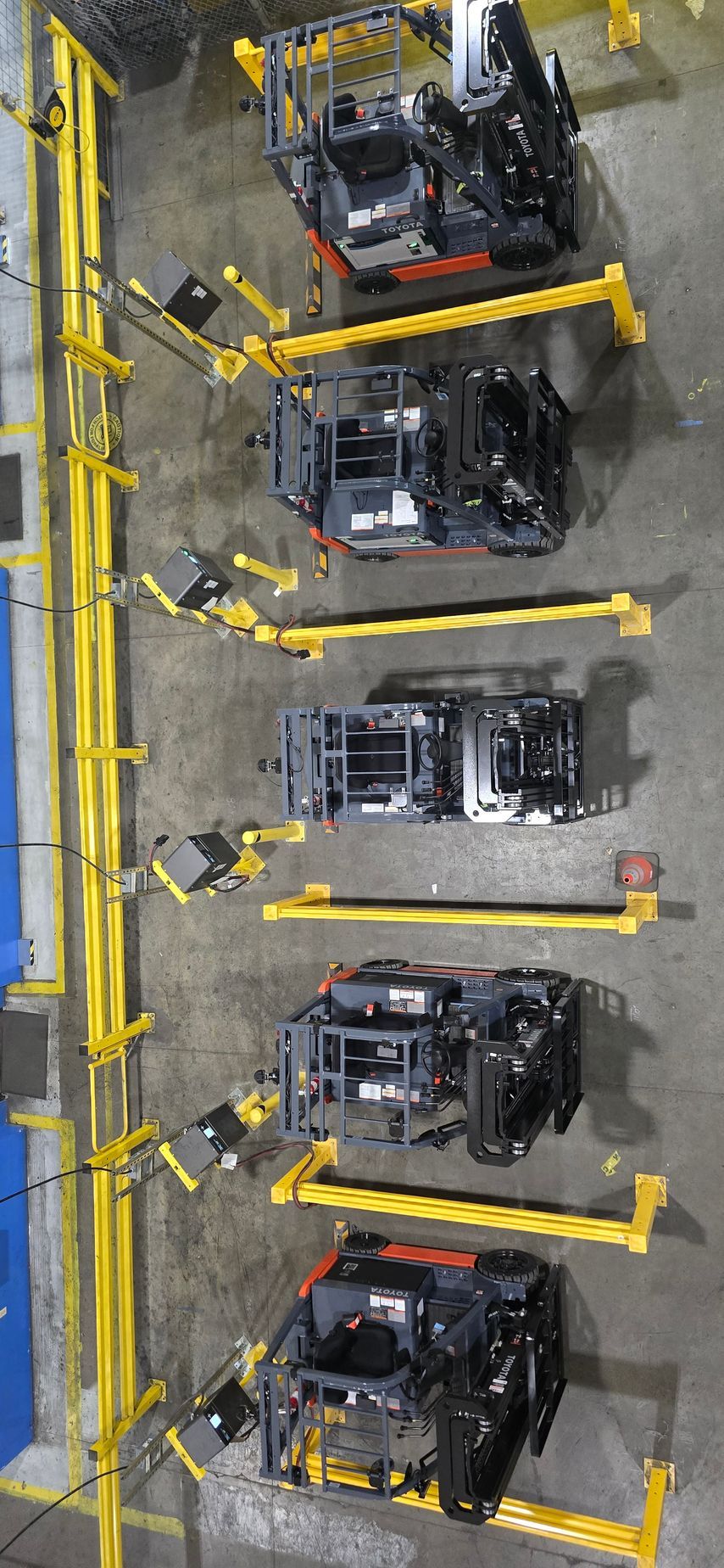 Five black forklifts parked in a warehouse rack, each secured behind yellow safety barriers.