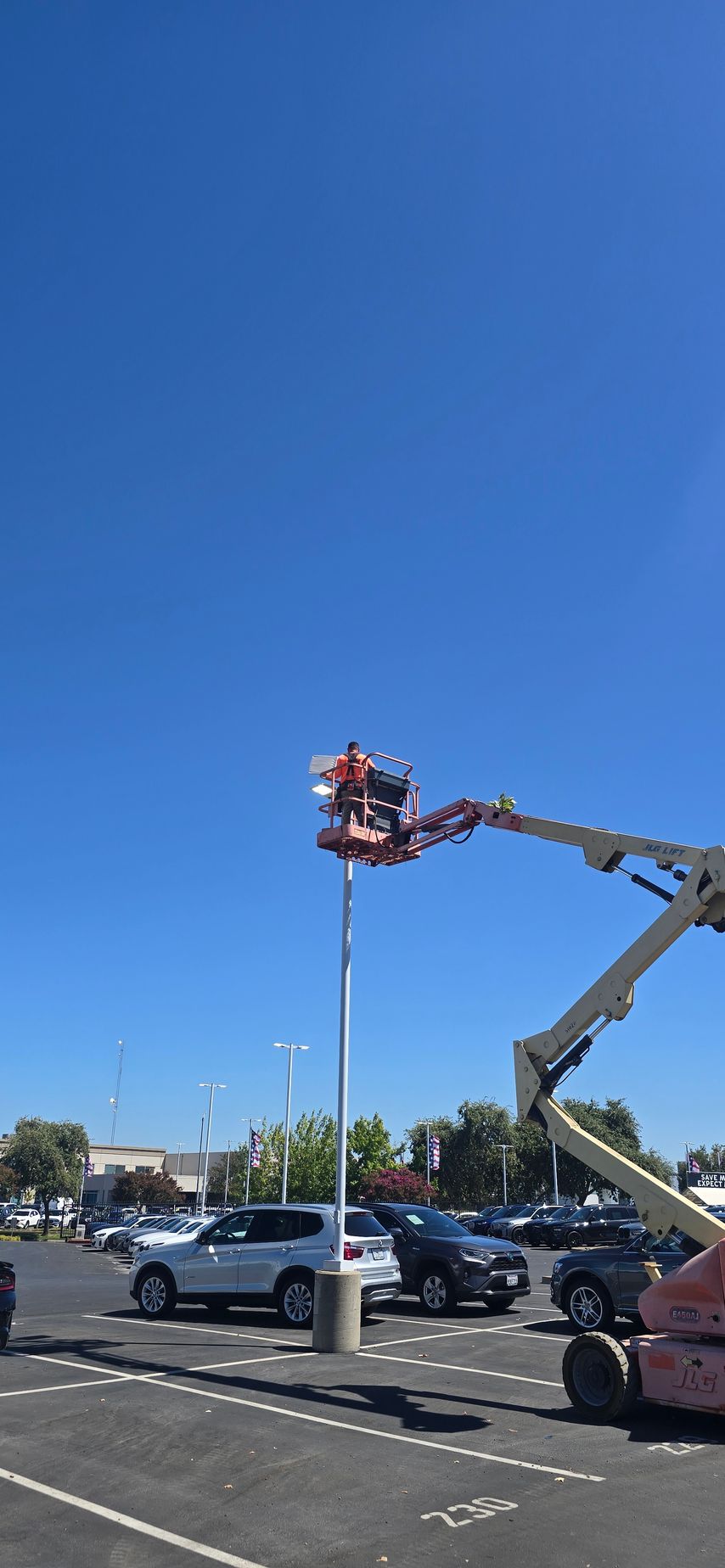Two workers in a red lift repairing a tall light pole in a parking lot under a clear blue sky