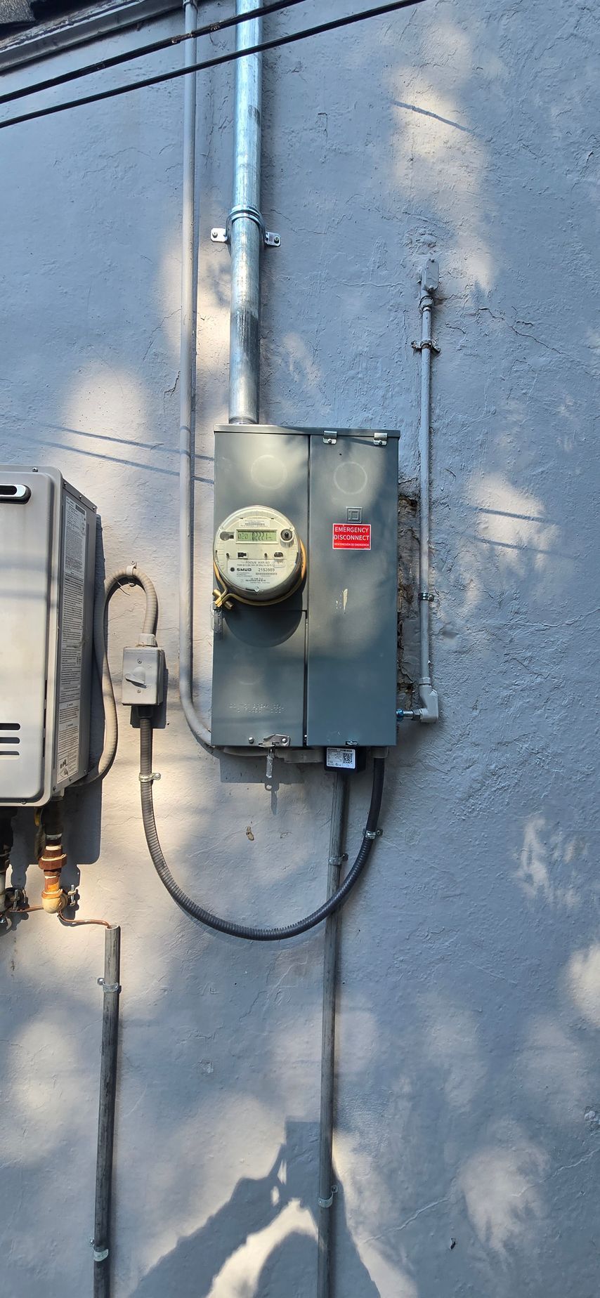 Gray electrical meter box mounted on a blue exterior wall with pipes and cables.