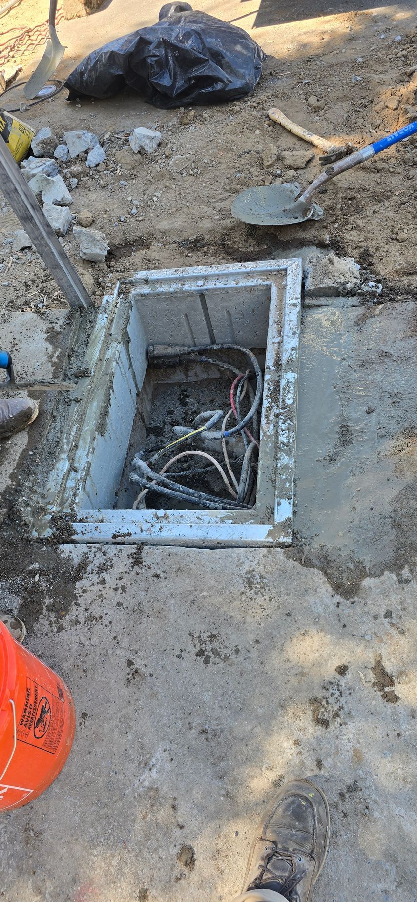 Open utility box in dirt beside a blue-handled tool and black bag at a construction site