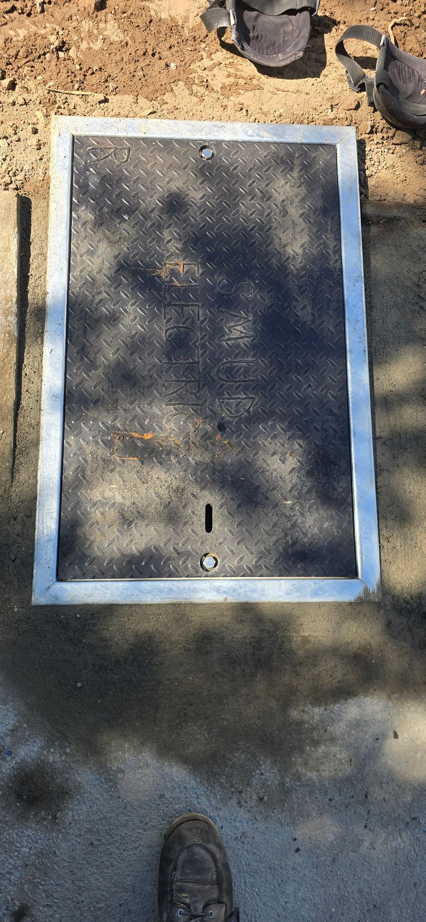 White-bordered rectangular utility cover on a concrete sidewalk, partly shaded by tree shadows.