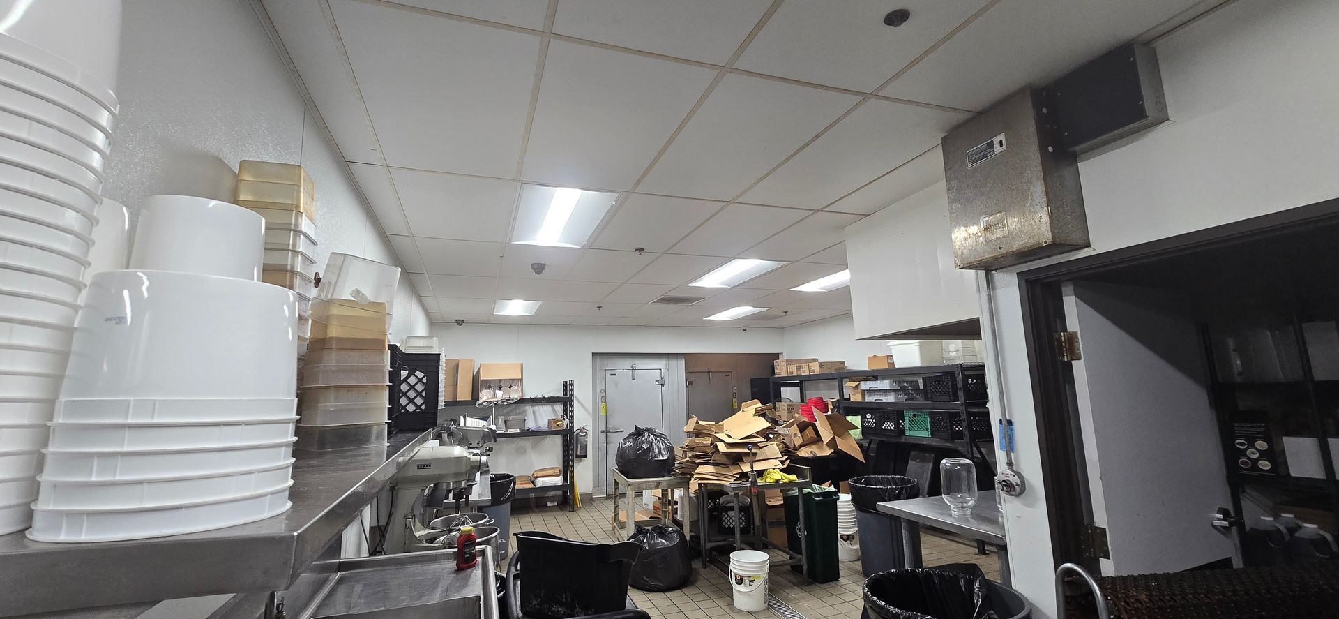 Cluttered commercial kitchen with stacked white containers, stainless counters, and a busy prep area
