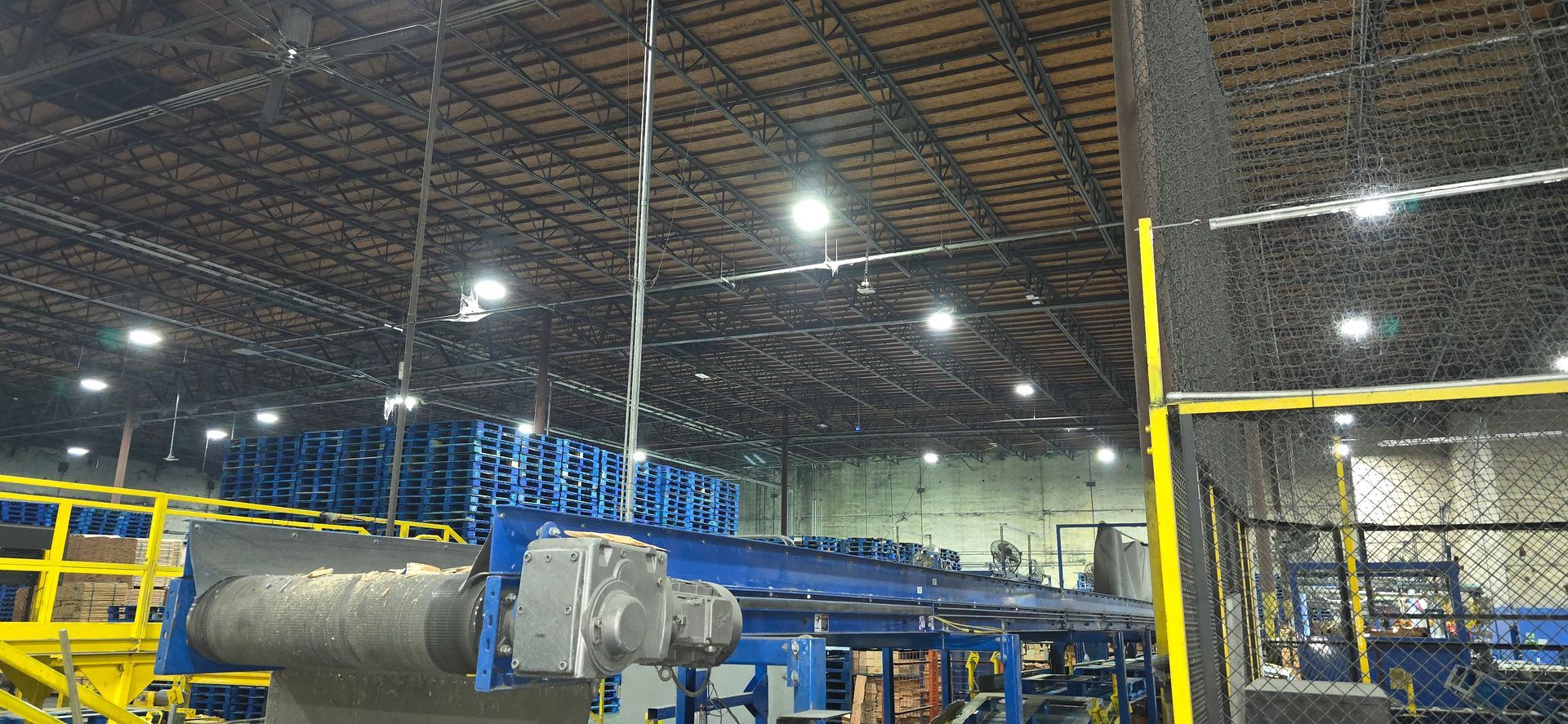 Industrial warehouse with blue conveyor machinery, yellow safety railings, and a large metal roll under bright lights