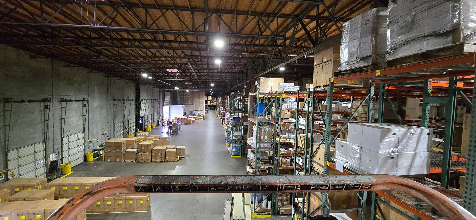 Wide warehouse interior with shelves, pallets, and a lit conveyor line under a high ceiling
