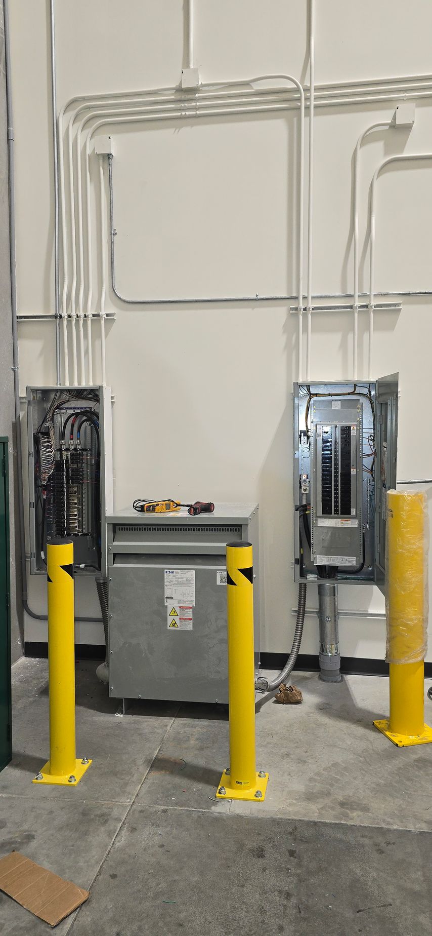 Industrial electrical panels and conduits on a wall, with yellow safety bollards in front.
