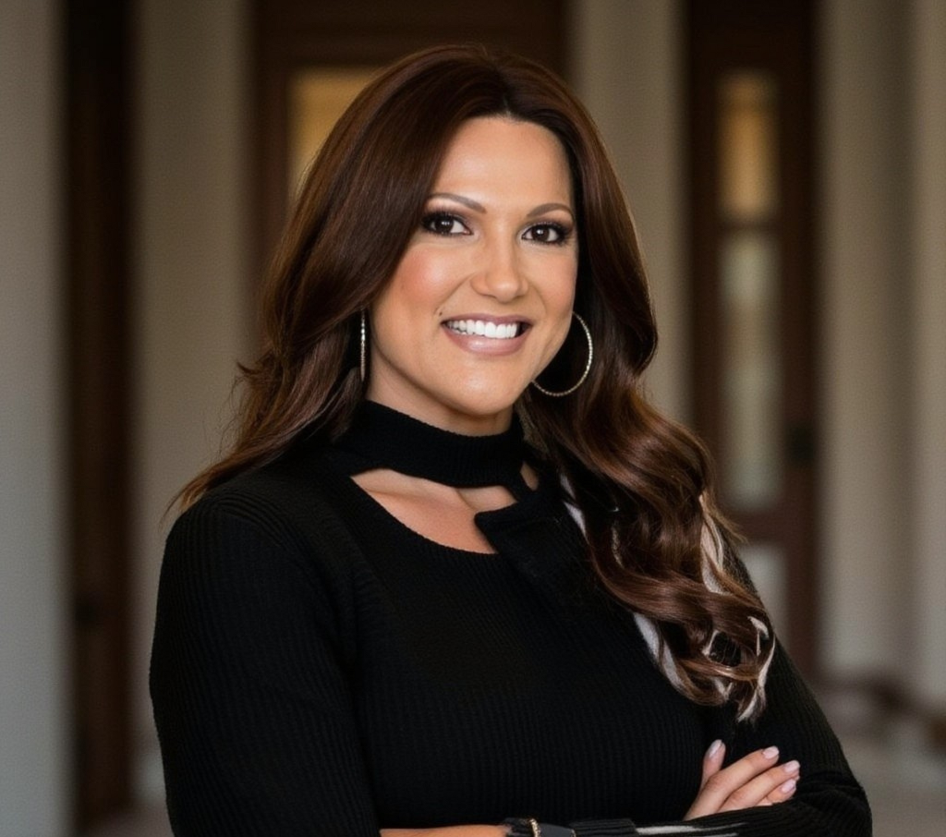 A person with long brown hair, wearing a black shirt with a cutout neckline and hoop earrings, smiles with arms crossed.