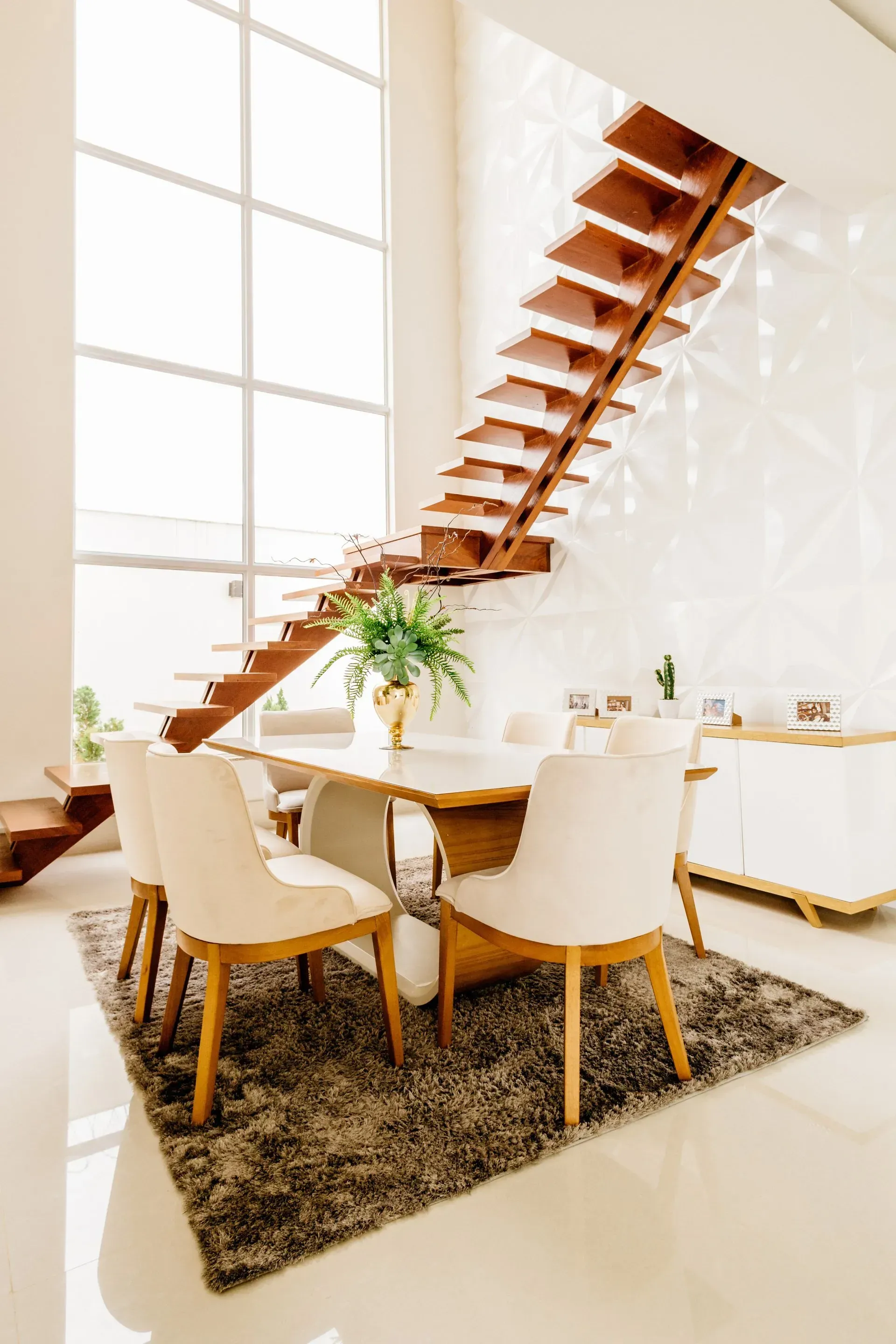 A dining area with four white chairs around a marble table, under a floating wooden staircase by a tall, grid-pane window.