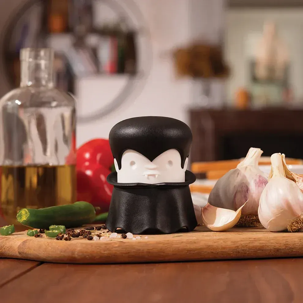 A black salt shaker with a face on it is sitting on a wooden cutting board surrounded by garlic and peppers.
