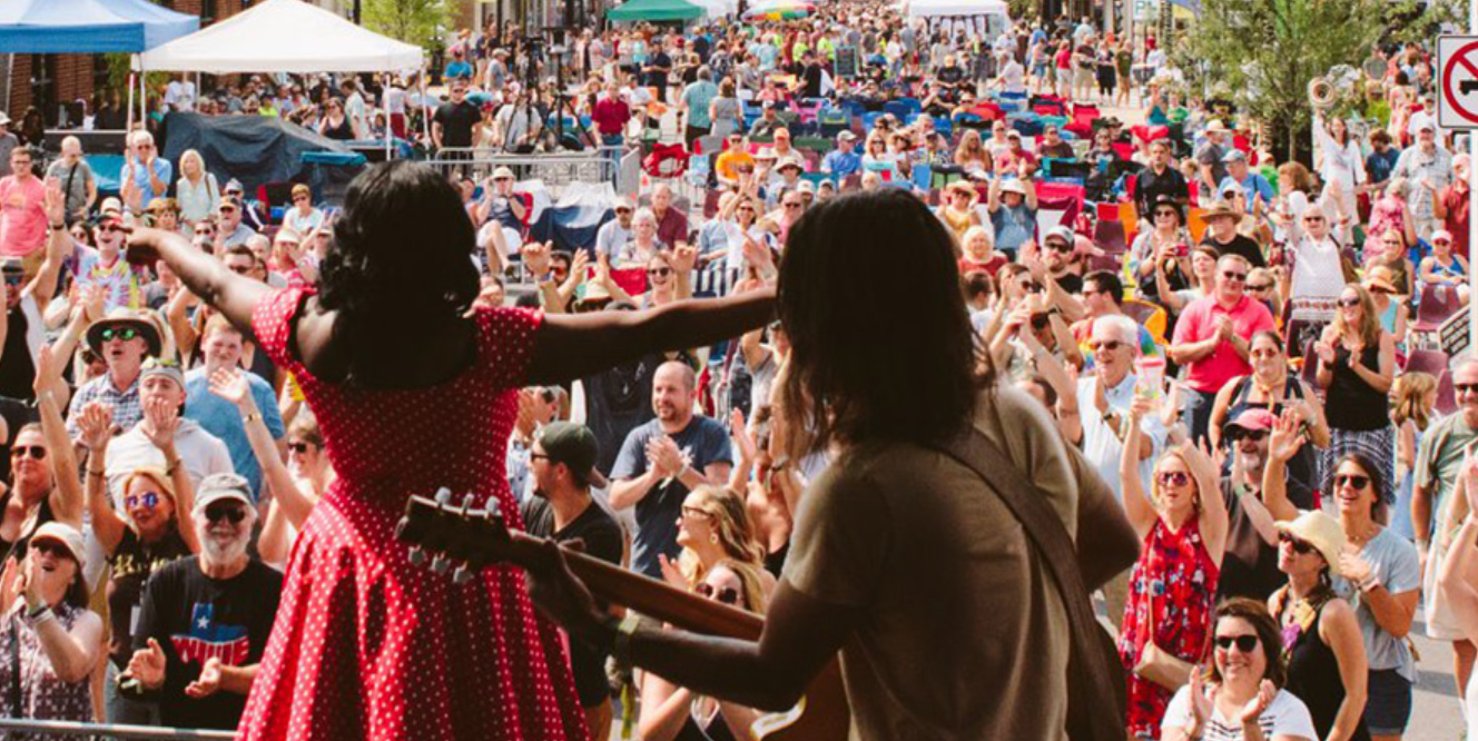 A woman in a red dress and a man with long hair are playing guitars and singing in front of a large crowd at the Rhythm & Roots Reunion Festival Stage in Bristol