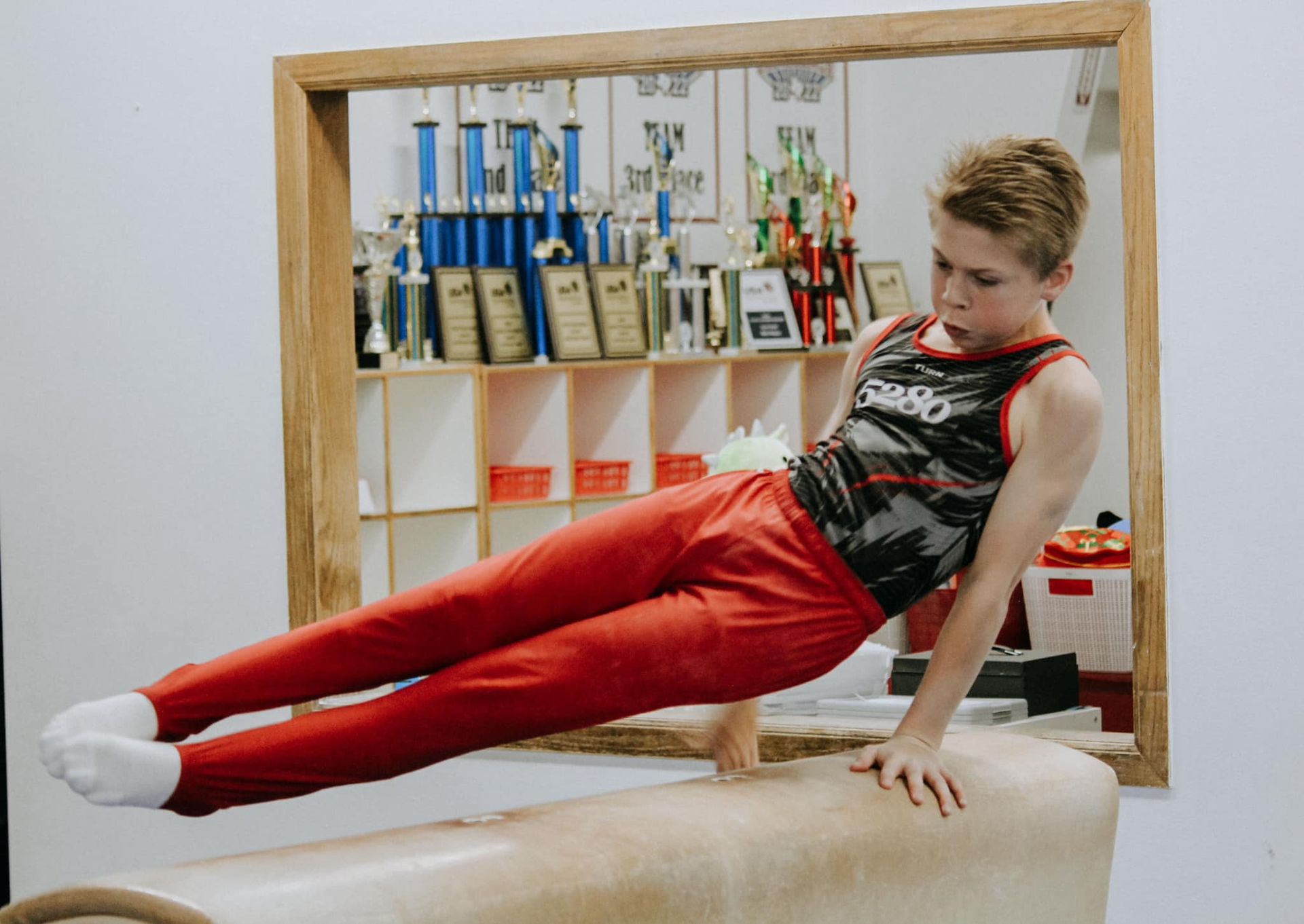 A young boy is doing a handstand on a pommel horse in front of a mirror.