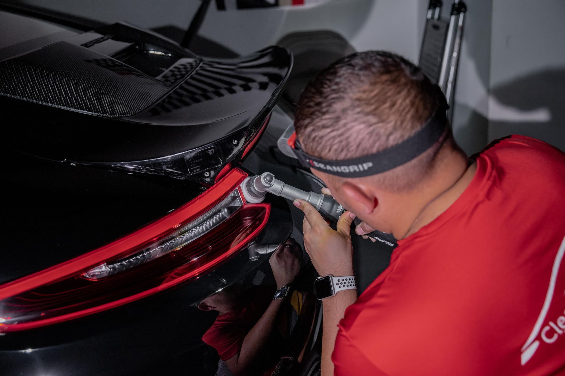 A man in a red shirt is polishing the back of a black car.